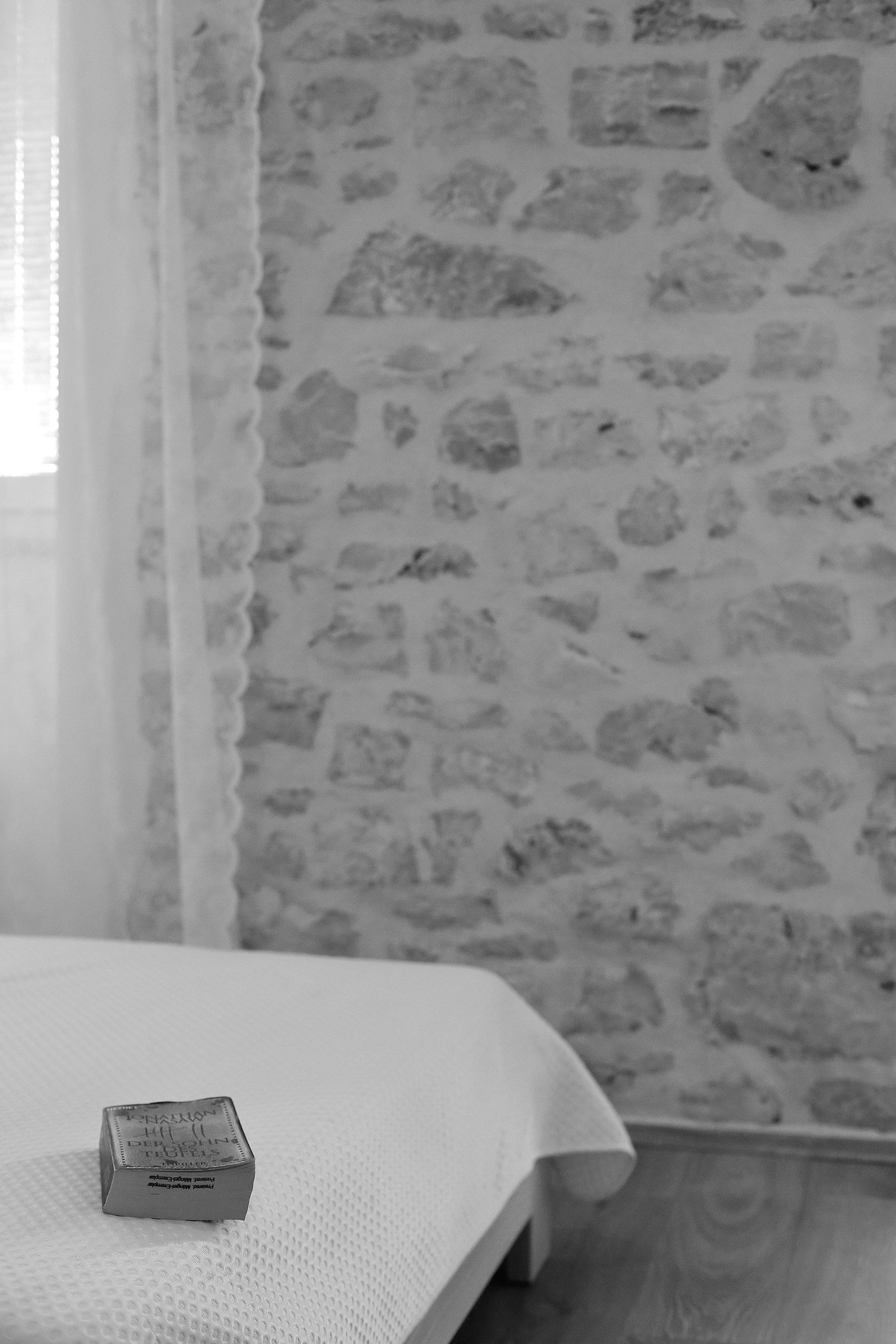 Una stanza con un letto, un piccolo scrigno sul letto, un muro in pietra e una finestra con tende leggere.