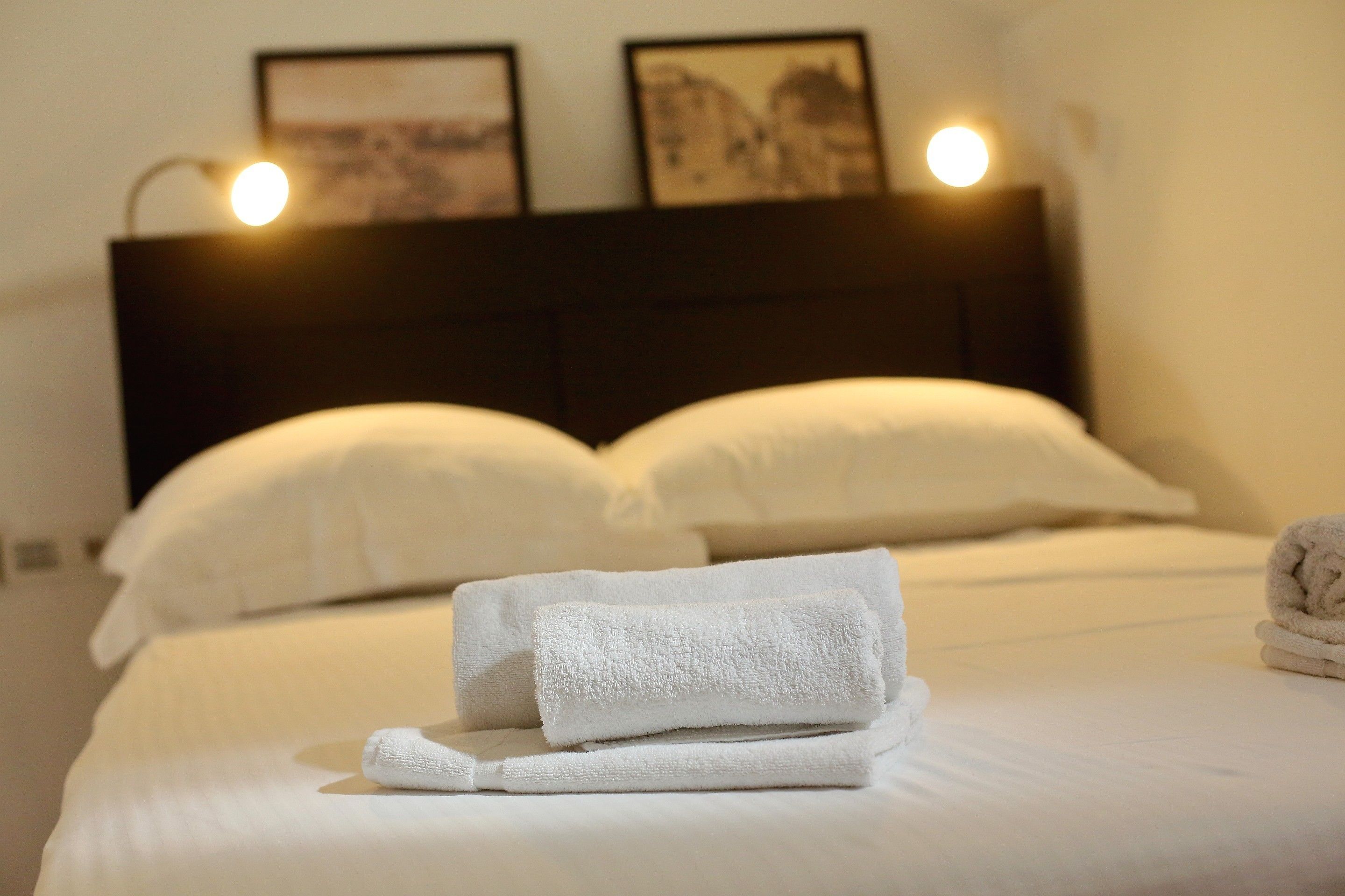 Cama bem arrumada com lençóis brancos e duas almofadas, uma toalha dobrada sobre a cama e duas fotografias emolduradas na parede acima da cama.