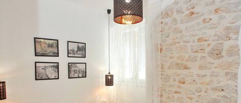 A bedroom with a wooden floor, a bed with white linens, a hanging light fixture, and a stone wall.