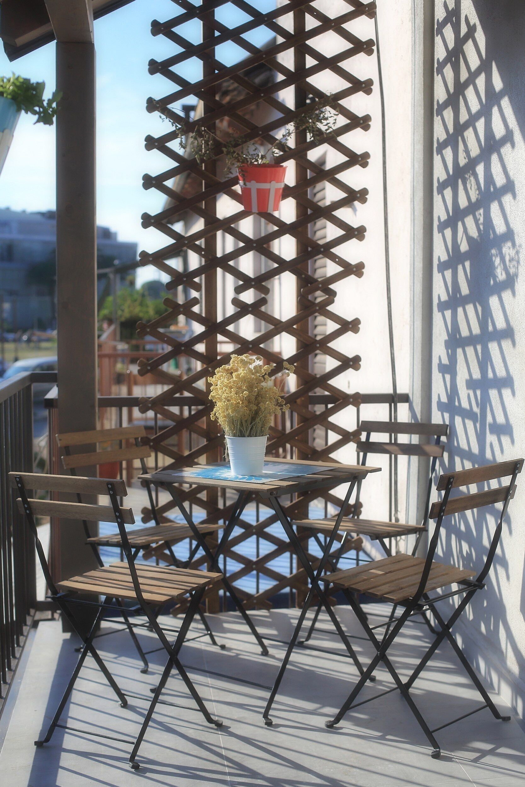 Uma varanda com mesa e cadeiras, um vaso de planta e uma estrutura treliçada.