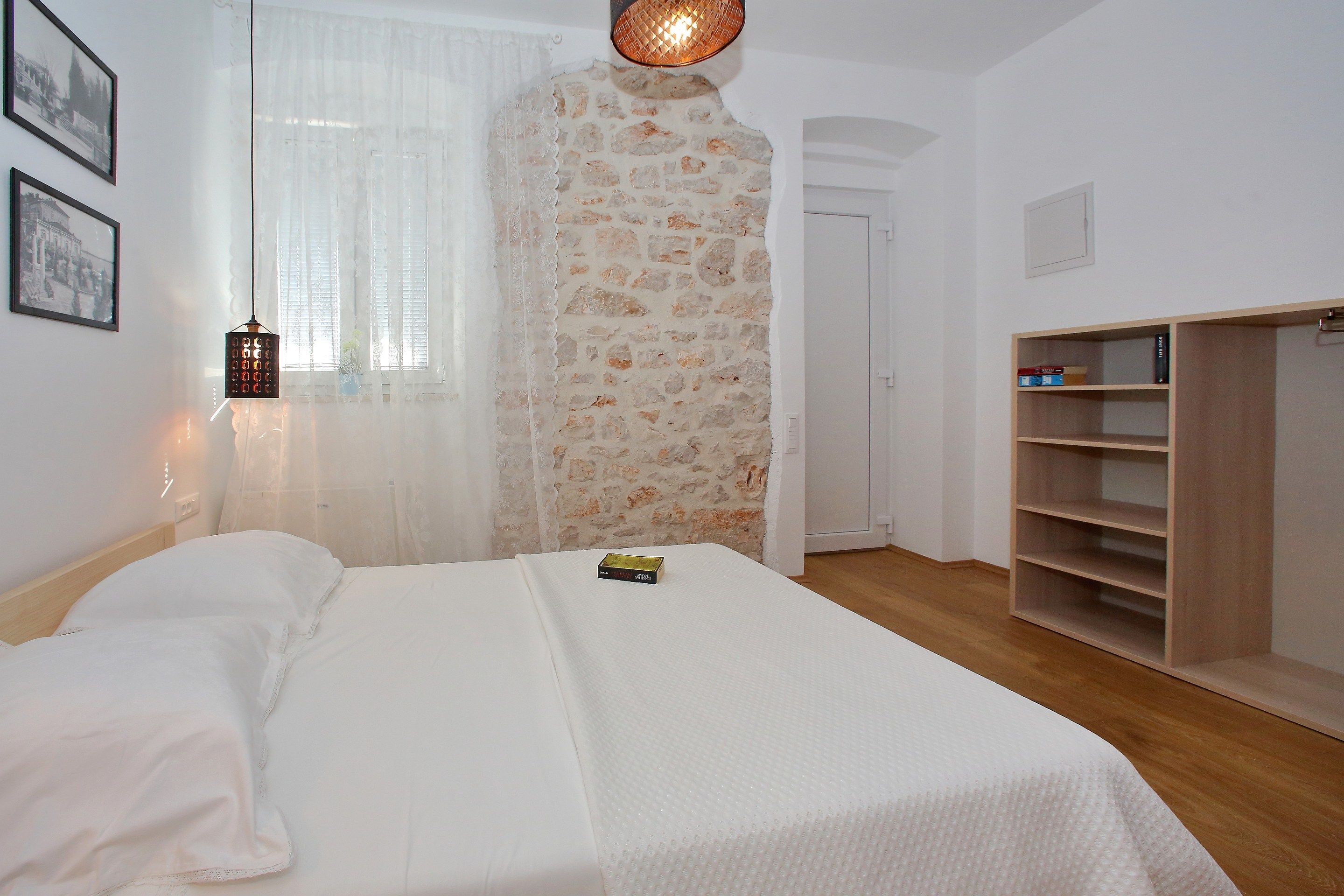 A bedroom with a bed, a wooden wardrobe, a stone wall, and a window with sheer curtains.