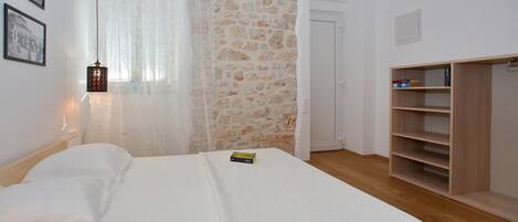 A bedroom with a bed, a wooden wardrobe, a stone wall, and a window with sheer curtains.