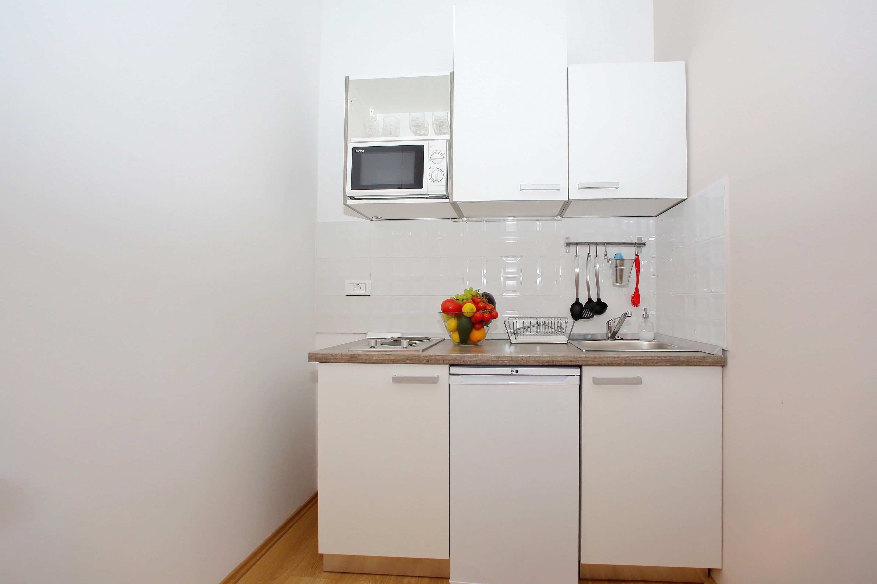 Una cocina compacta con muebles blancos, microondas, lavamanos y frutero.