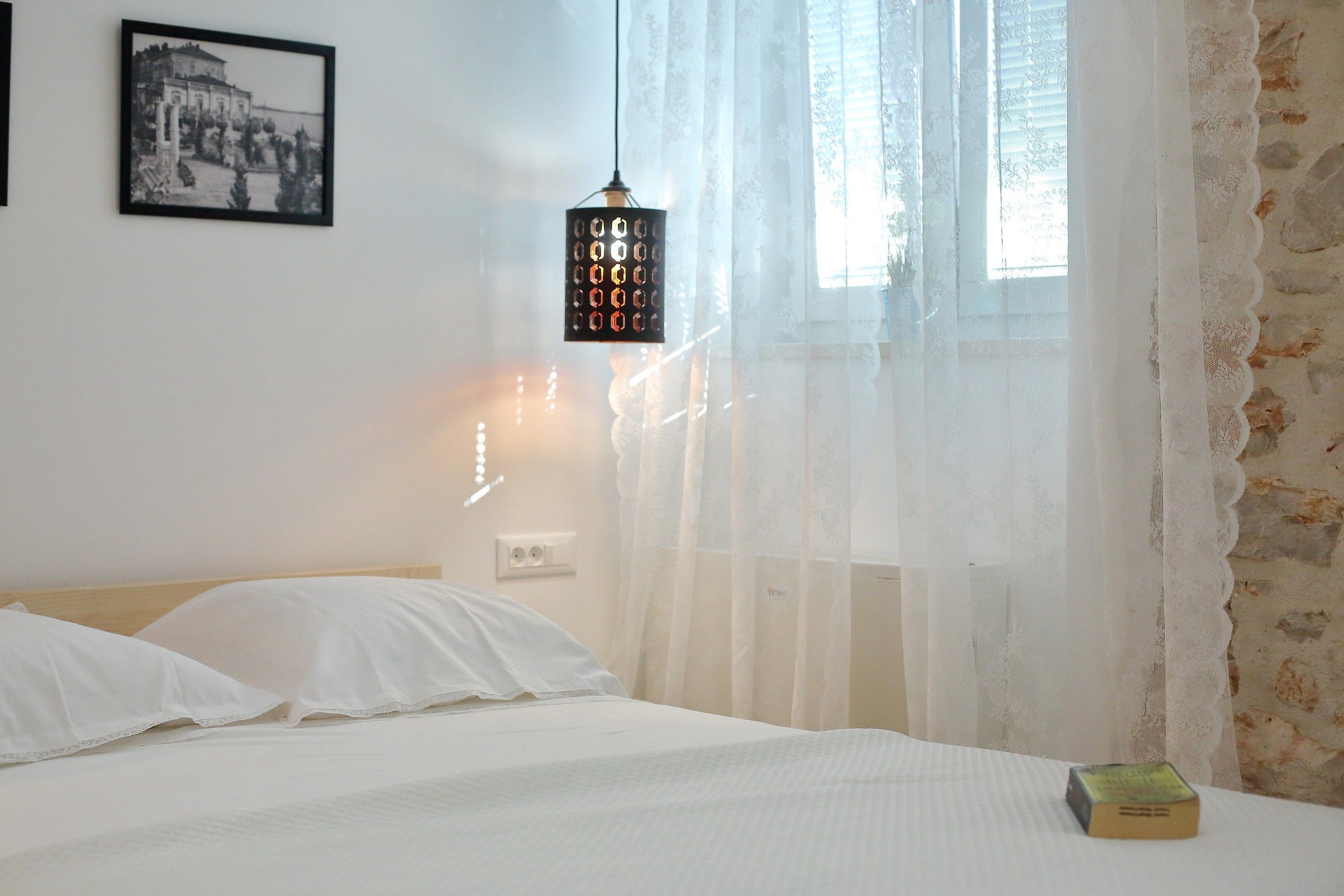 Un dormitorio con cama, ropa de cama blanca, lámpara colgante, un cuadro enmarcado en la pared y una ventana con cortinas traslúcidas.