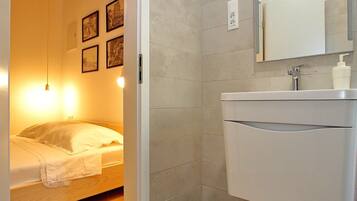 A compact bedroom with a wooden ceiling, a bed with white bedding, a bedside table, and a wall-mounted light fixture. Adjacent is a bathroom with a white sink, mirror, and a shower area.