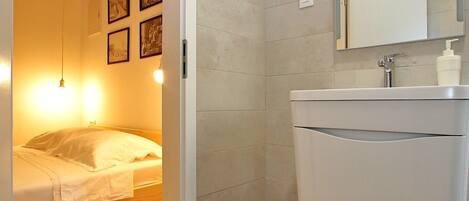 A compact bedroom with a wooden ceiling, a bed with white bedding, a bedside table, and a wall-mounted light fixture. Adjacent is a bathroom with a white sink, mirror, and a shower area.