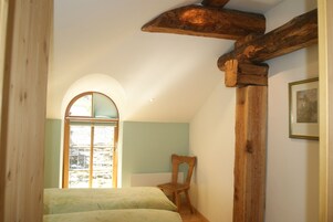 Un dormitorio con viga de techo de madera, una cama con colcha verde estampada, una silla de madera y una ventana con vistas al exterior.