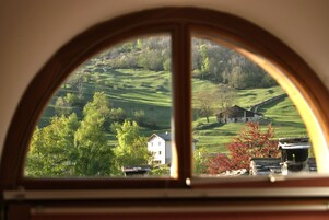Vista a través de una ventana con marco de madera que muestra un paisaje rural con colinas onduladas, árboles y una casa blanca.