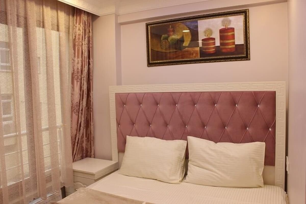 A bedroom with a tufted headboard, two pillows, a bedside table, and a window with sheer curtains.