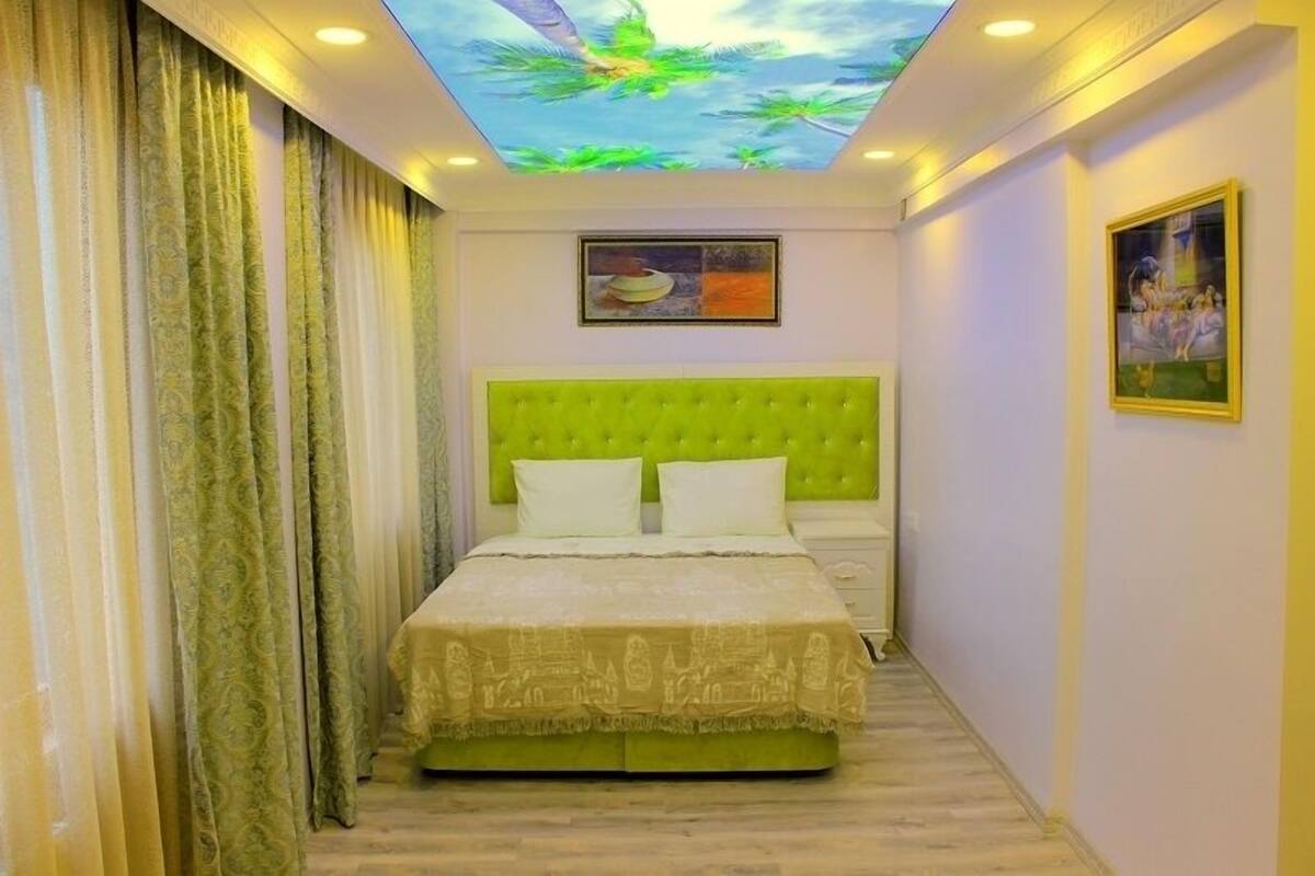 A bedroom with a green upholstered headboard, a bed with white linens, a ceiling with a tropical mural, and two framed artworks on the walls.