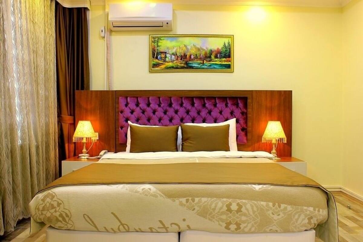 A hotel room with a large bed, two bedside lamps, a painting on the wall, and an air conditioning unit.
