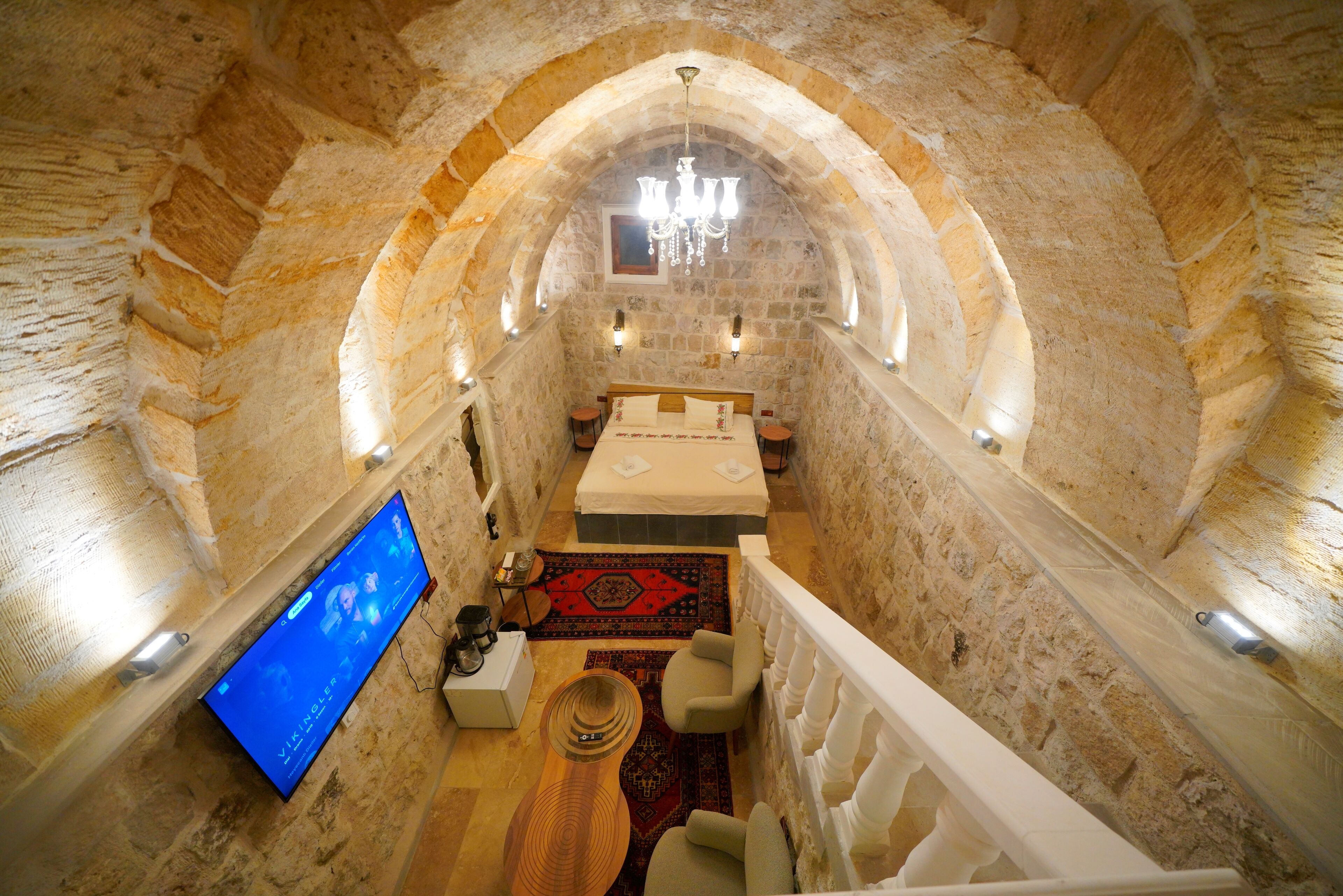 A narrow stone-walled room with a bed, a flat-screen TV, a wooden table, and a red patterned rug.