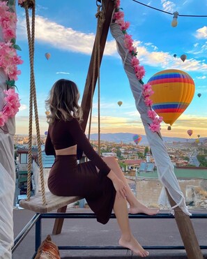 A woman sitting on a swing with ropes, overlooking a cityscape with hot air balloons in the sky. - View Cave Hotel (Nevsehir)