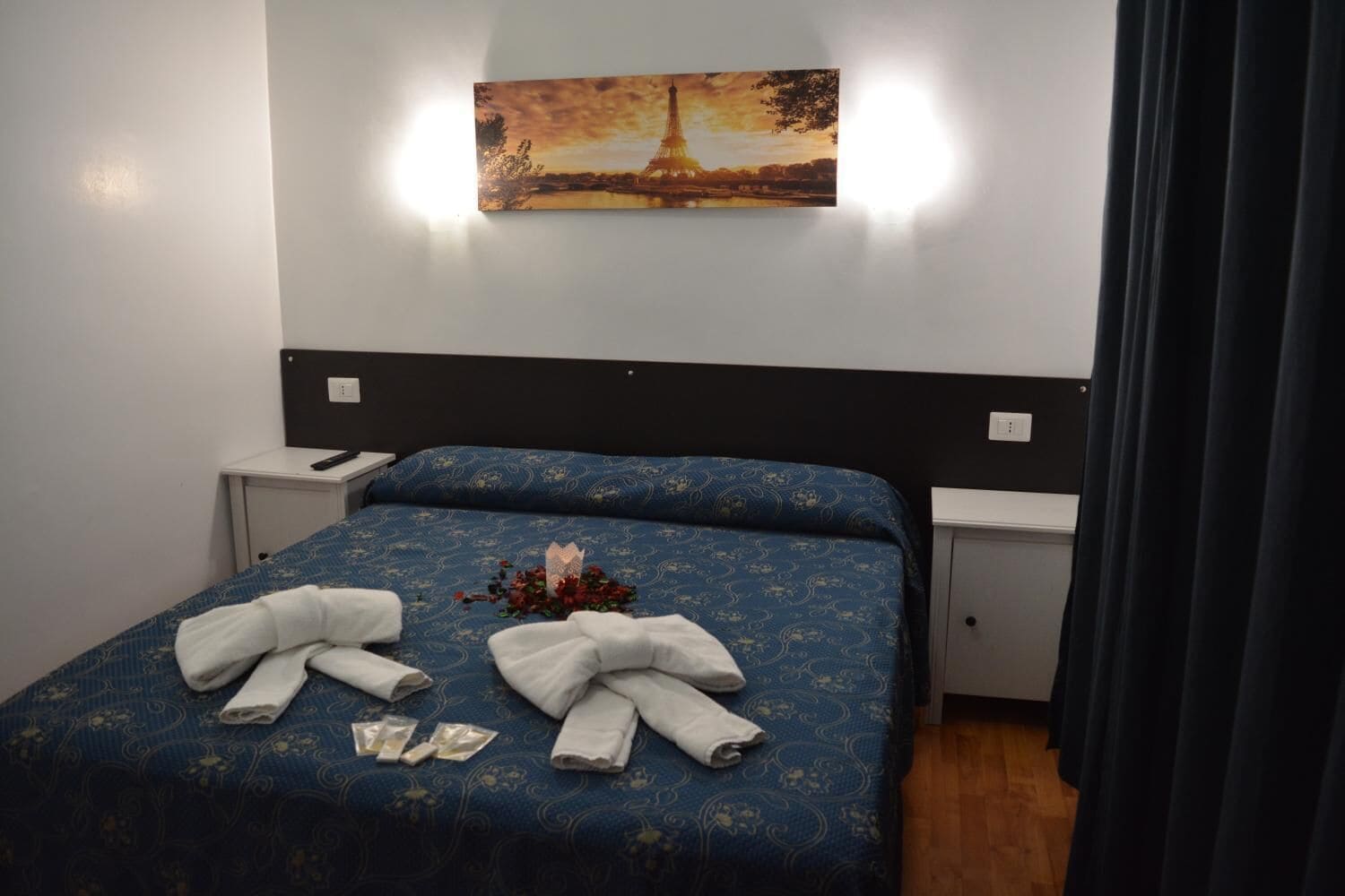 A neatly made bed with a blue patterned bedspread and white towels, a painting of the Eiffel Tower on the wall, and a small bedside table with a lamp.