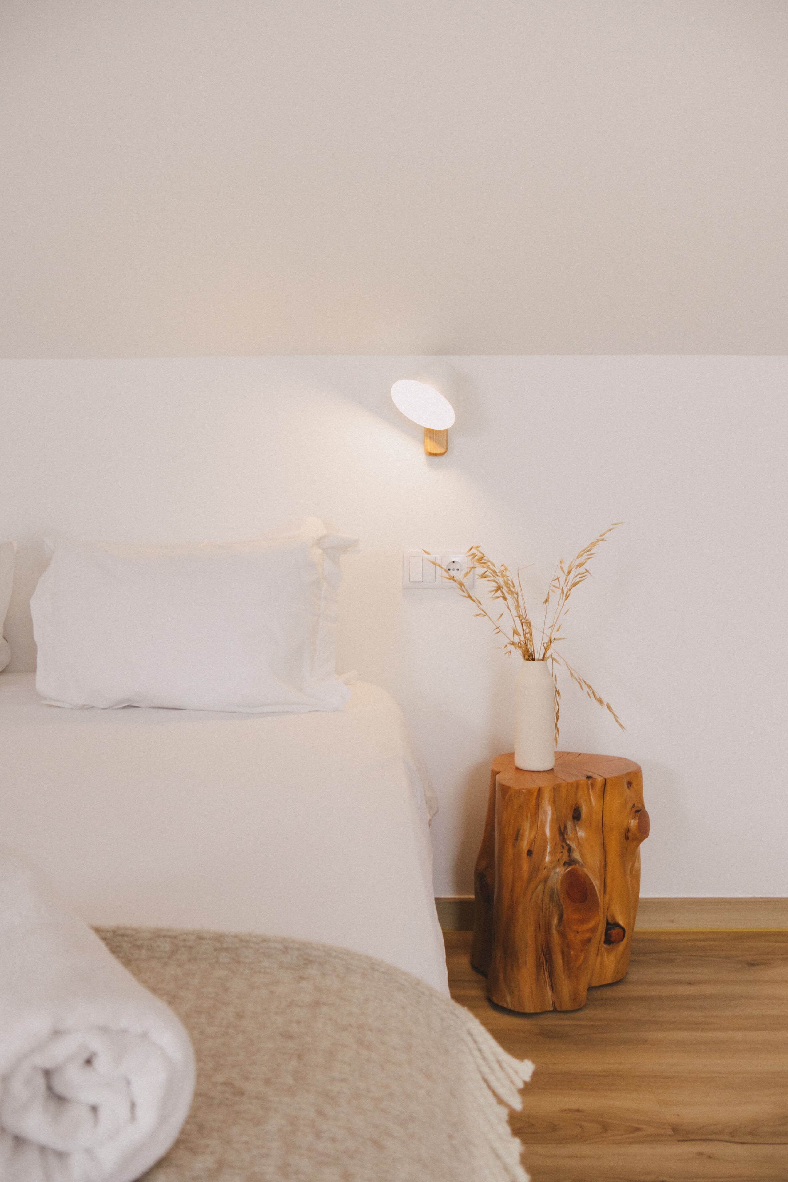 A neatly made bed with white bedding, a wooden nightstand with a vase of dried plants, and a wall-mounted light fixture.
