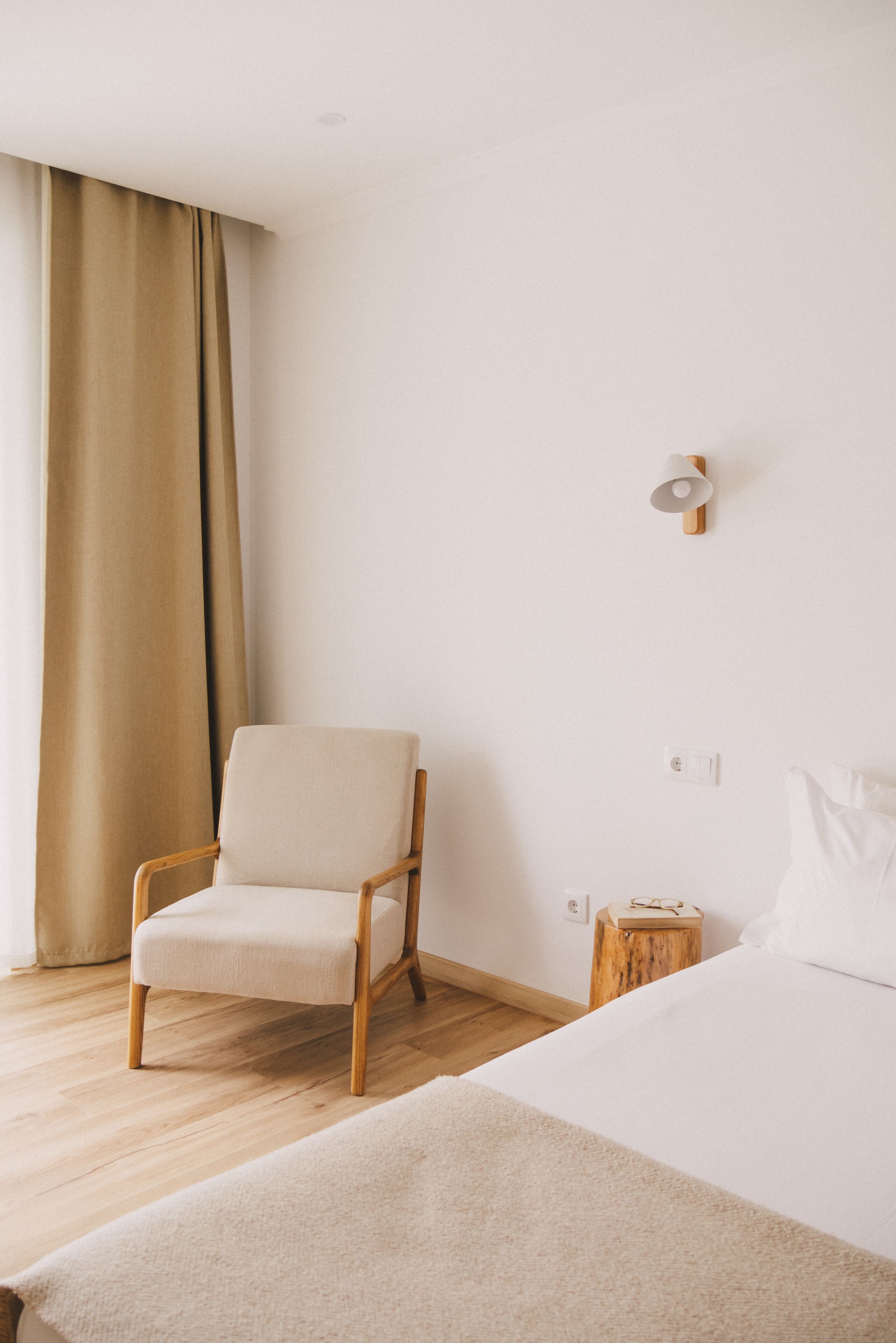 Camera d'albergo dal design minimalista, con pavimento in legno, una sedia beige, un letto bianco e un piccolo comodino in legno.