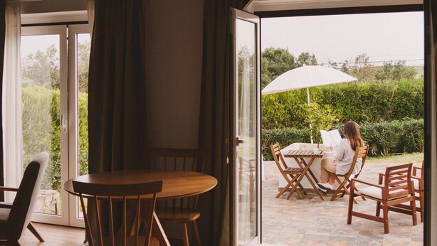 A wooden dining table with chairs inside a room with a glass door leading to an outdoor patio area. - Pure Flor de Esteva (Vila do Bispo)