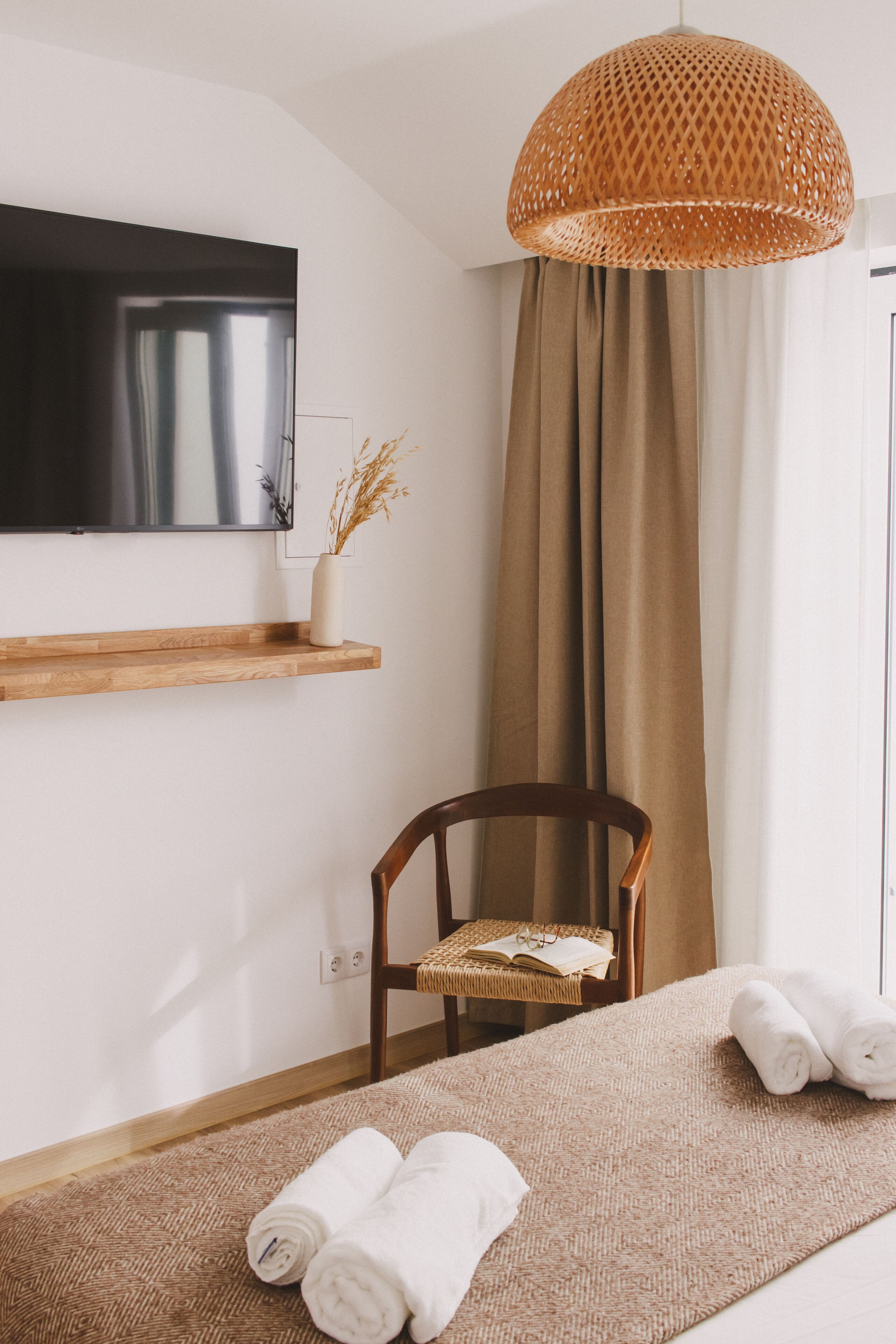 Une chambre d’hôtel avec un lit, une chaise en bois, un téléviseur fixé au mur, un luminaire en osier et une fenêtre avec des rideaux.