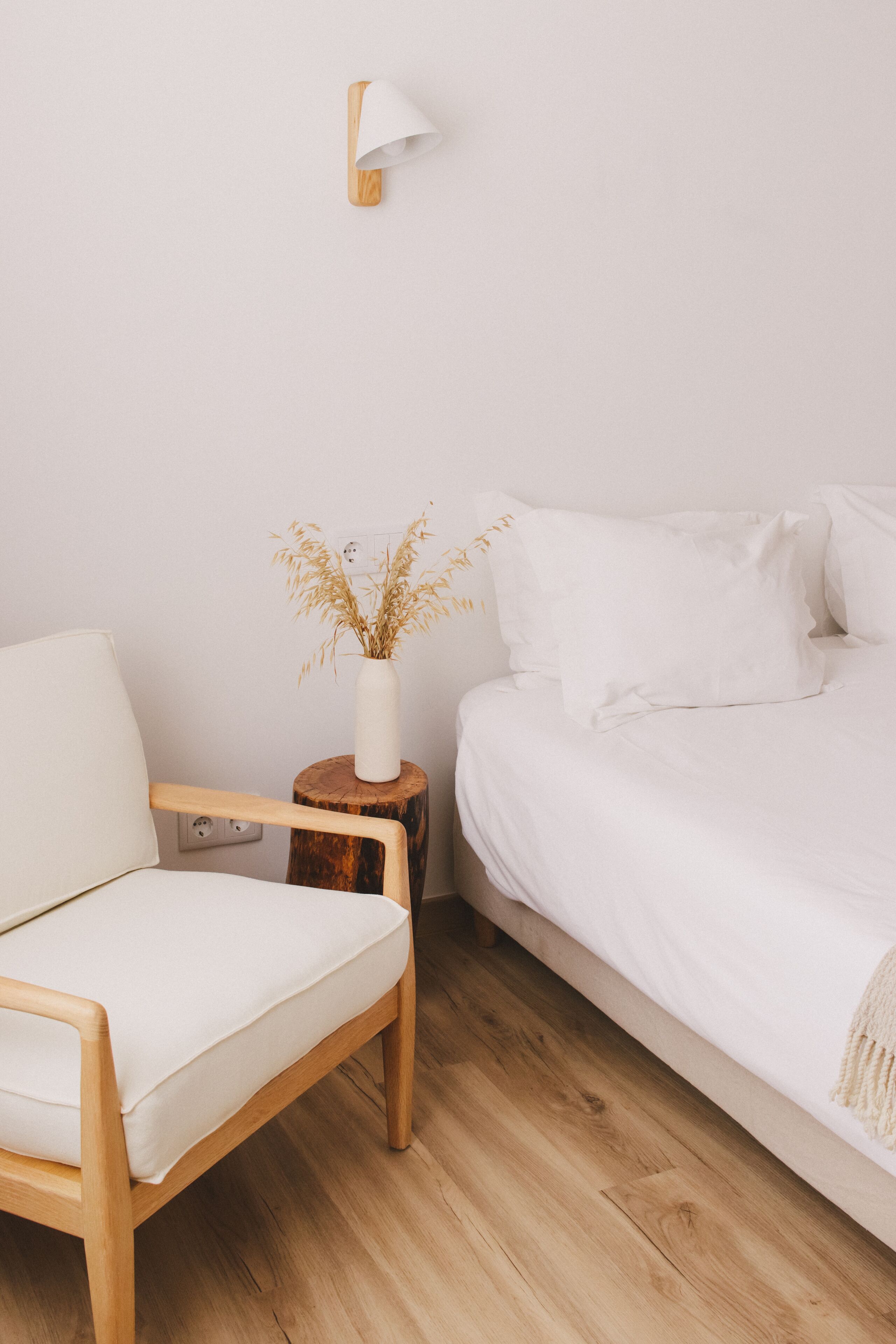 A minimalist bedroom with a wooden floor, a white bed, a white chair, and a bedside table with a vase of dried plants.