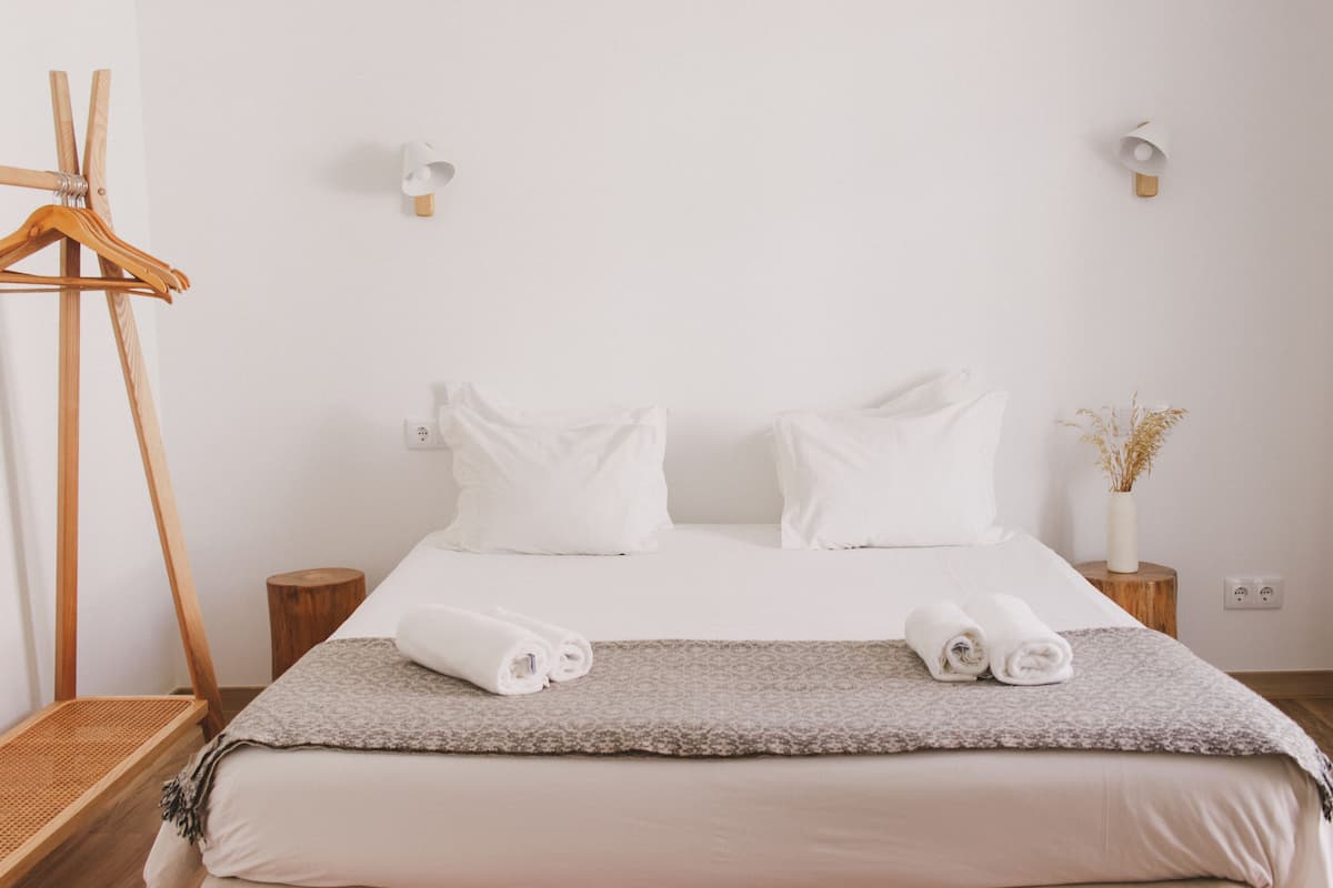A neatly made bed with white linens and two rolled towels, a wooden hanger on the left, and a small wooden nightstand with a vase of dried flowers.