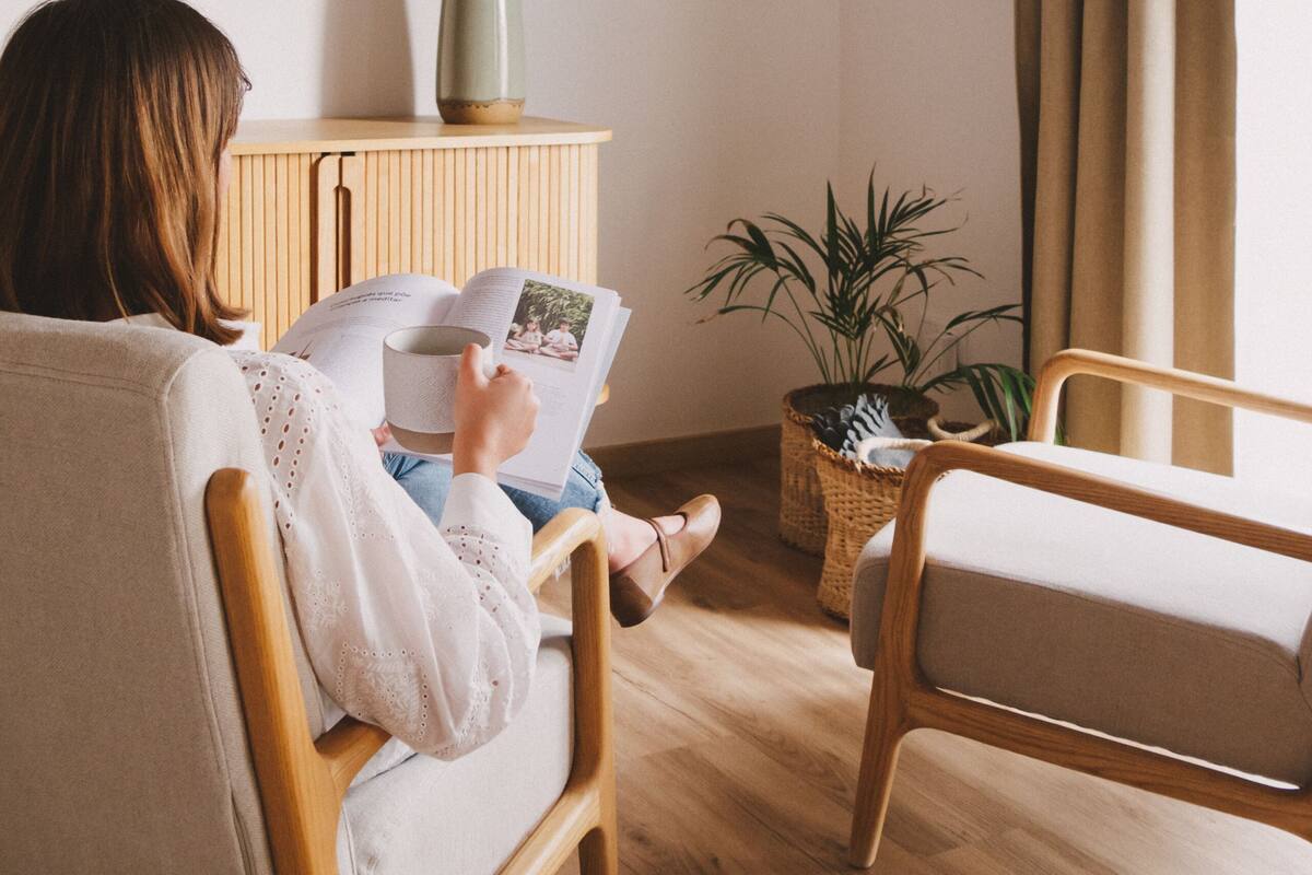 A person reading a book in a room with a wooden floor, a television mounted on the wall, a potted plant, and a window with curtains.