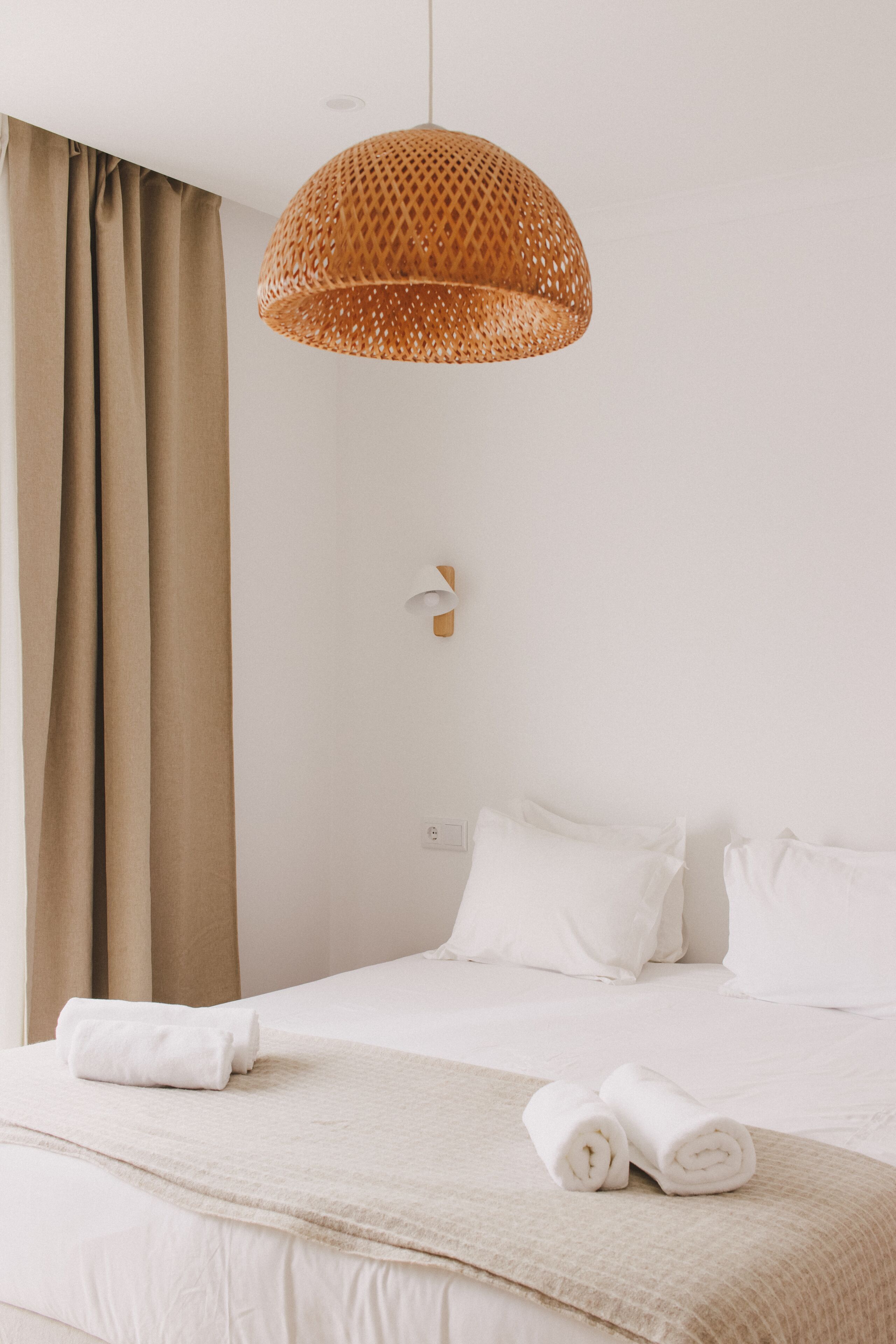 A hotel room with a bed, beige curtains, a woven pendant light, and white bedding.