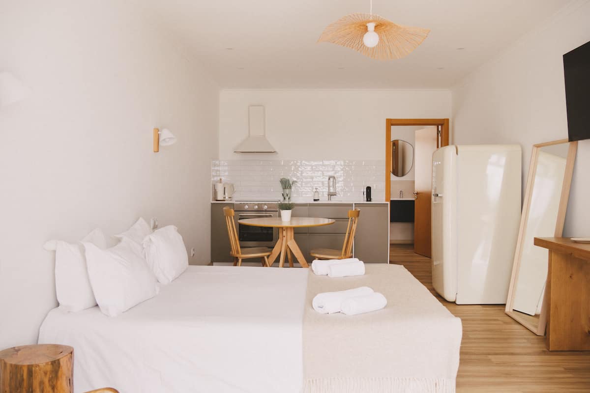 A modern hotel room with a large bed, a wooden side table, a round dining table with chairs, a kitchen area with a stove, and a refrigerator.