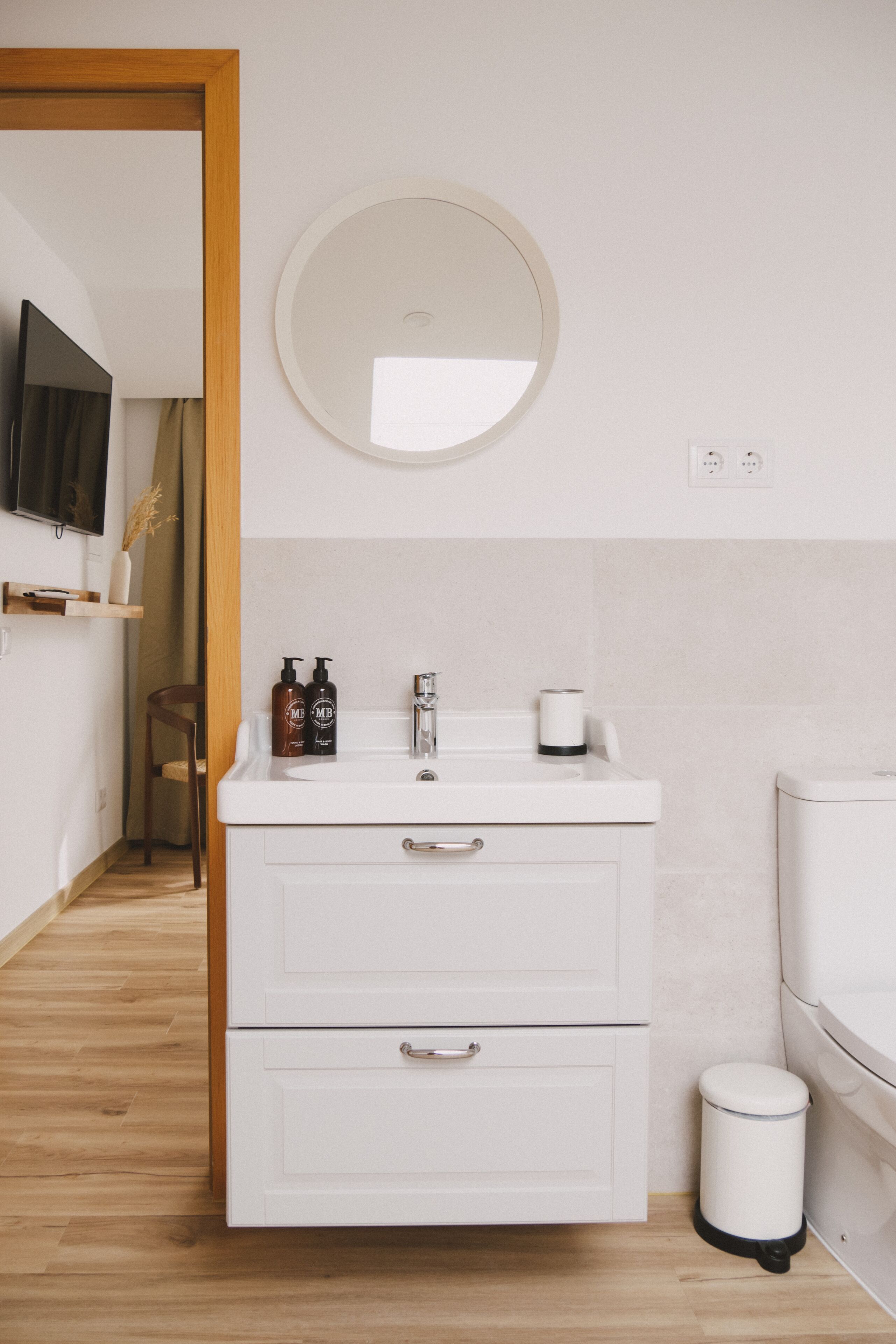 Ein modernes Badezimmer mit einem weißen Waschtisch, einem runden Spiegel und einer kleinen Mülleimer.