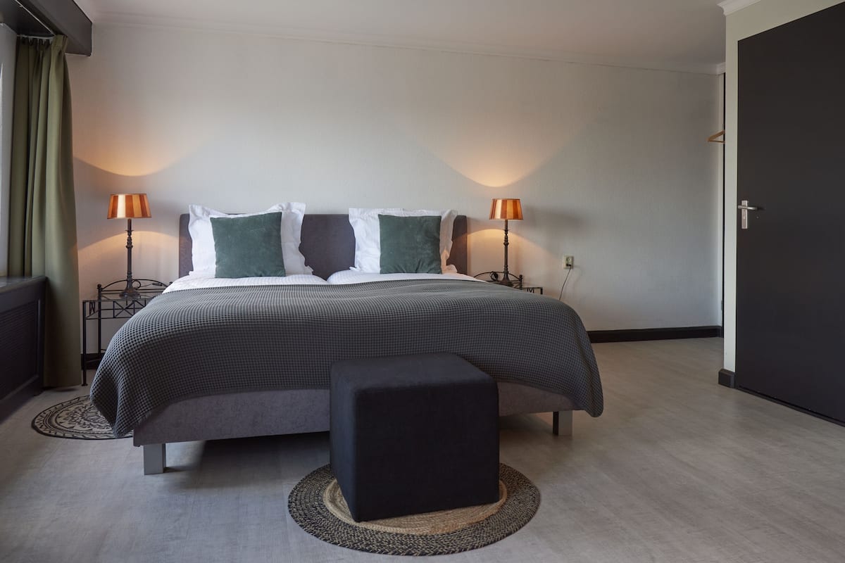A modern bedroom with a large bed, two bedside lamps, a small ottoman, and a dark door.