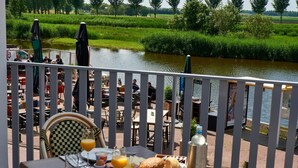 A dining area with tables and chairs overlooking a river, with a view of lush greenery and a distant field. - Hotel Villa GROET uit Avenhorn (Avenhorn)
