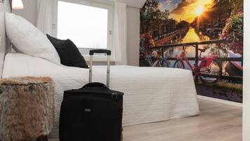 Une chambre avec un lit, une valise, un petit tabouret moelleux et une fresque murale représentant un paysage de canal.