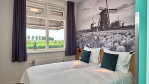 A double bed with white bedding and green pillows, a window with curtains, and a mural of windmills and a field. - Hotel Villa GROET uit Avenhorn (Avenhorn)