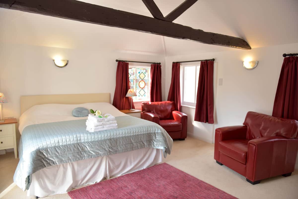 A bedroom with a large bed, two red leather chairs, a window with red curtains, and a wall-mounted lamp.