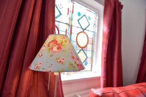 Una habitación con una lámpara floral, cortinas rojas y una ventana de vitral.