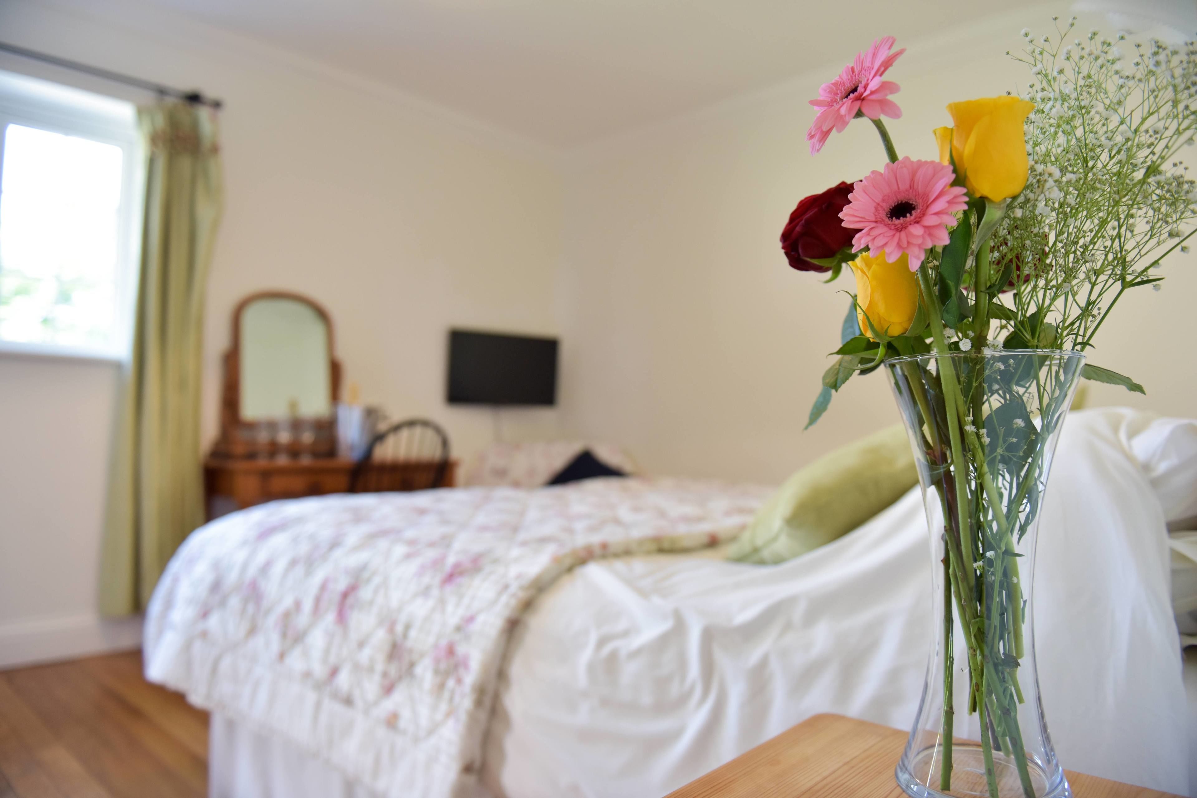 Un dormitorio con cama, cómoda, espejo y un florero con flores sobre una mesa.