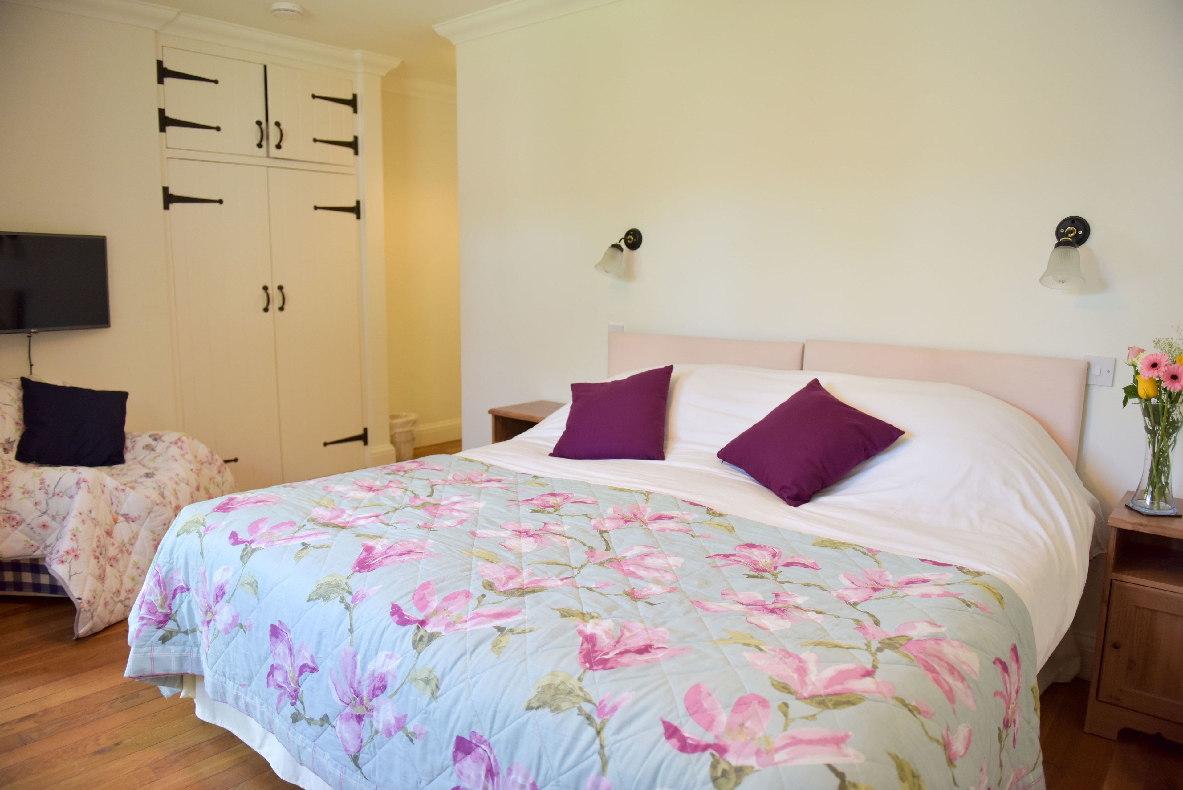 A bedroom with a floral bedspread, two pillows, a nightstand, a TV, and a wardrobe.