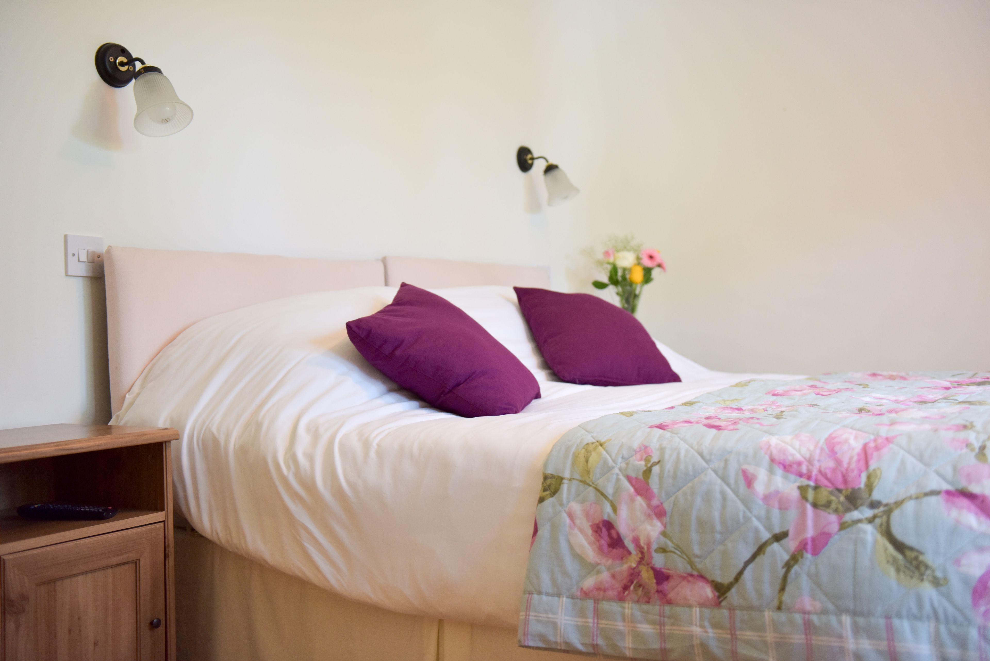 Una cama bien hecha con edredón floral, una mesita de noche de madera y una lámpara de pared.
