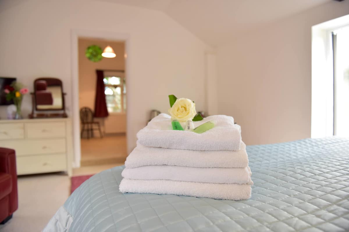 A neatly made bed with a quilted blue cover, a stack of white towels, and a single white rose on top.