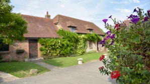 Un bâtiment en briques avec un toit en pente, un chemin menant à l’entrée et un jardin agrémenté de fleurs violettes.