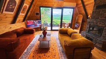 A cozy living room with a wooden ceiling, a stone fireplace, and a large window offering a view of the outdoors.