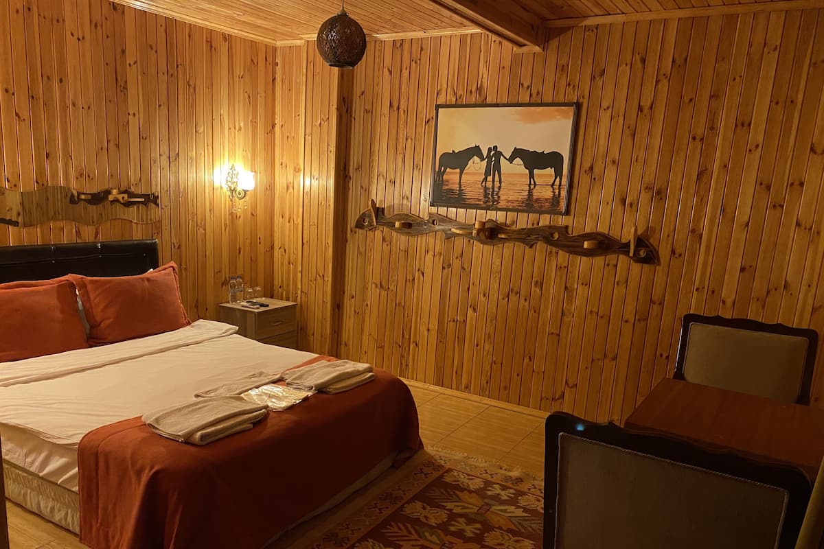 A wooden-paneled room with a bed, a chair, a nightstand, a lamp, and a painting of horses.