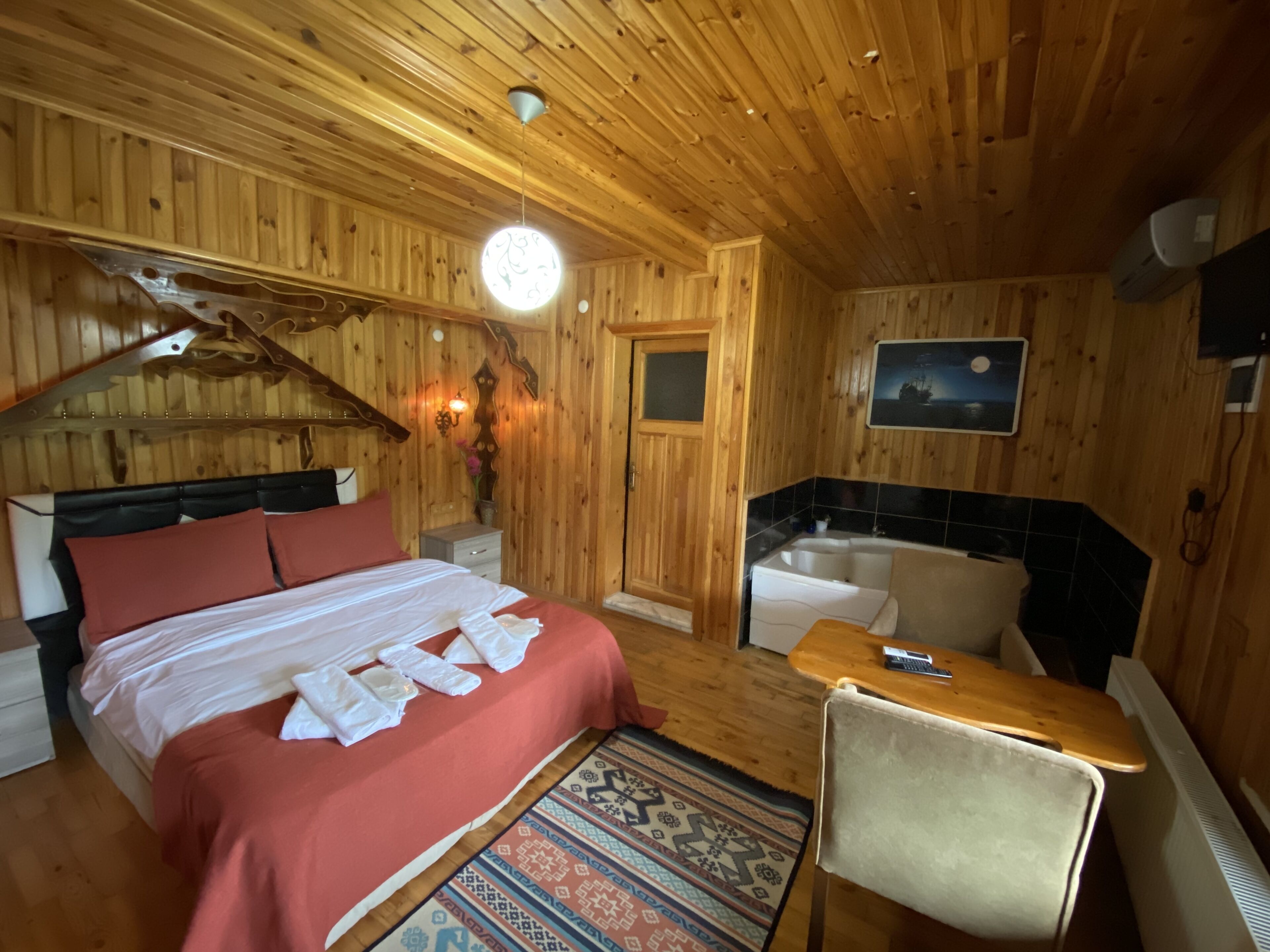 A wooden cabin room with a bed, a chair, a small table, a sink, and a TV.