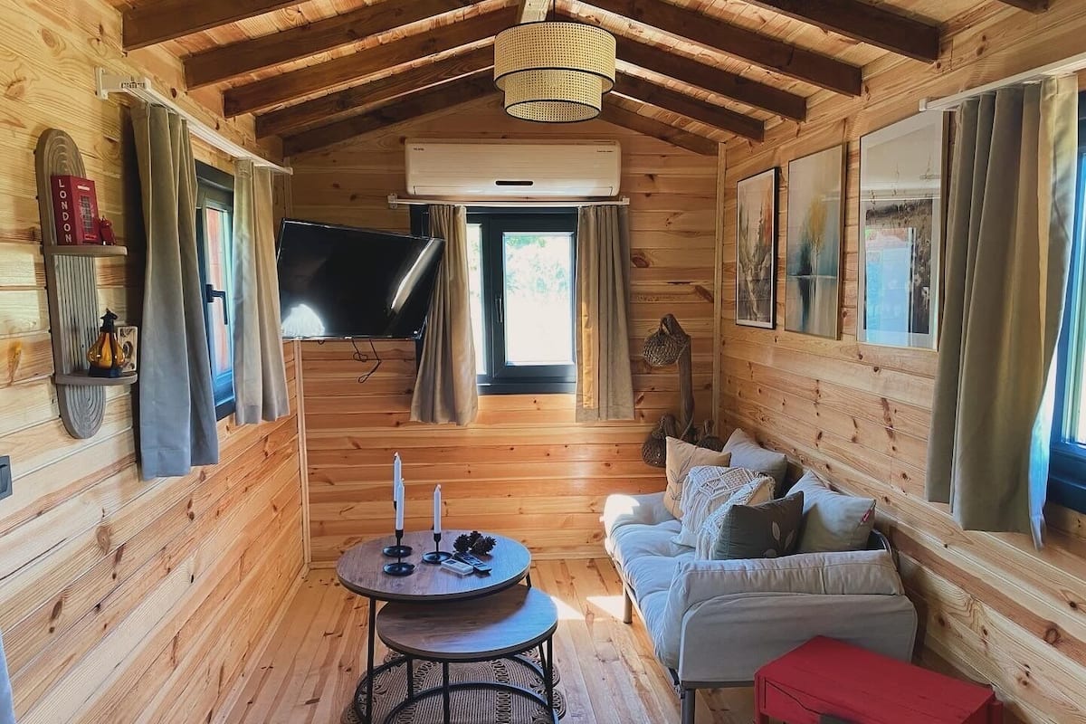 A cozy cabin interior with a wooden interior, a sofa, a red bench, a round table with candles, and a mounted TV.