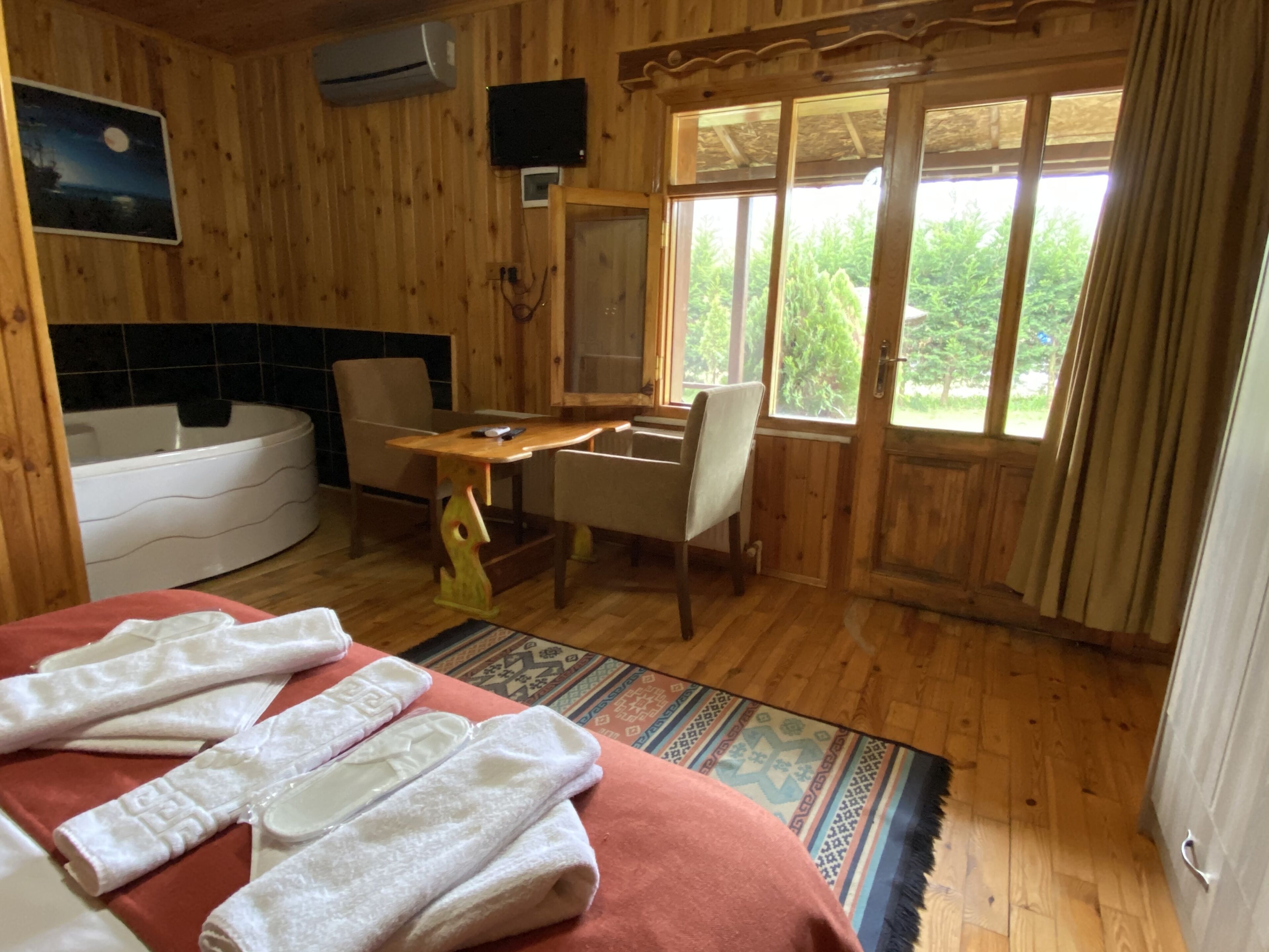 A wooden cabin room with a bed, a desk, a chair, a bathtub, and a large window with a view of greenery.