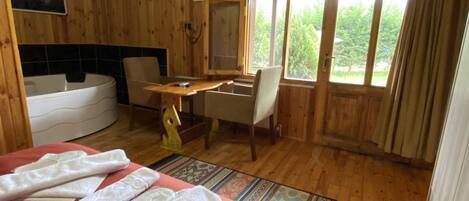 A wooden cabin room with a bed, a desk, a chair, a bathtub, and a large window with a view of greenery.