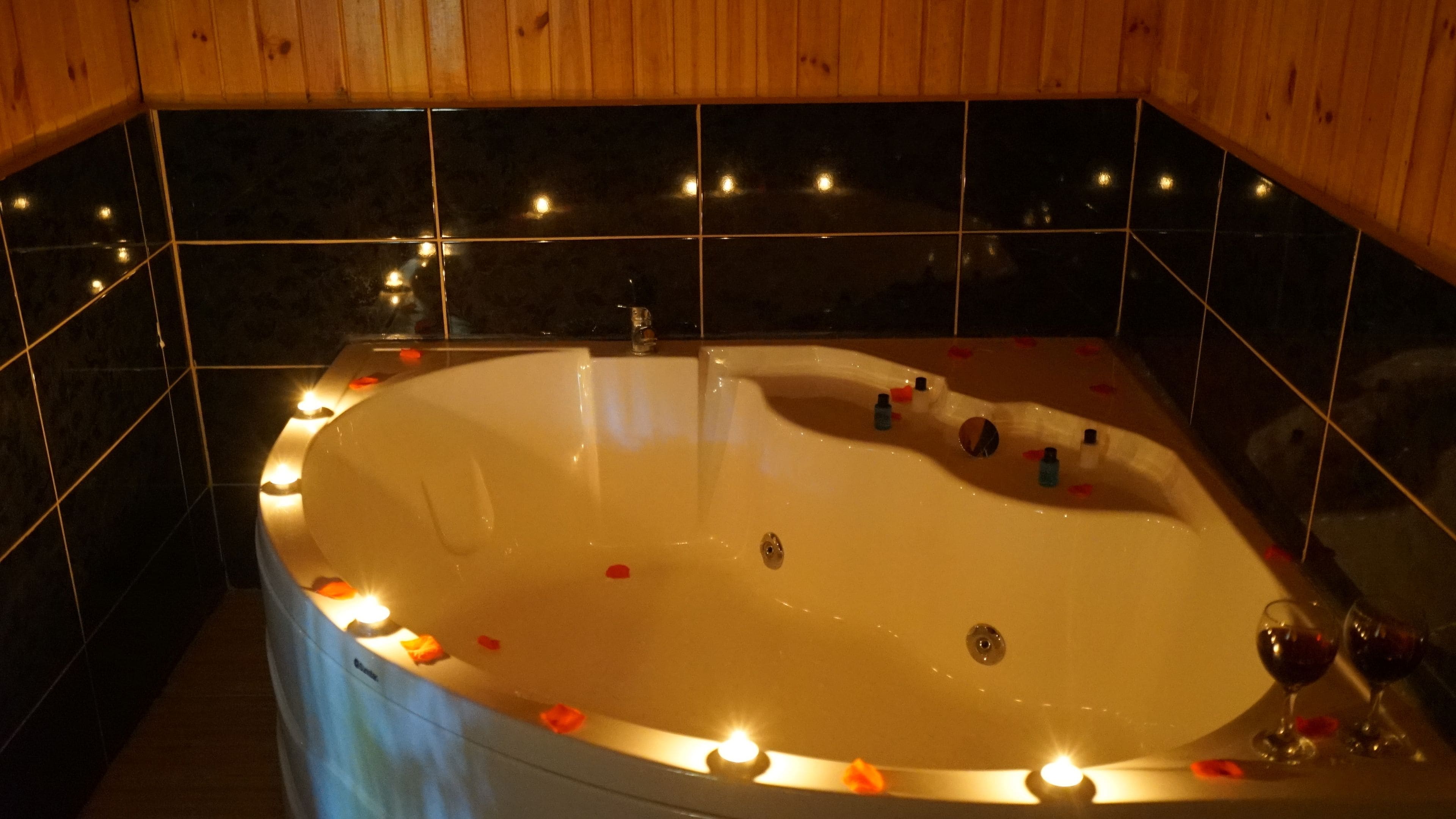 A bathtub with rose petals and candles, surrounded by dark tiles and wooden paneling.