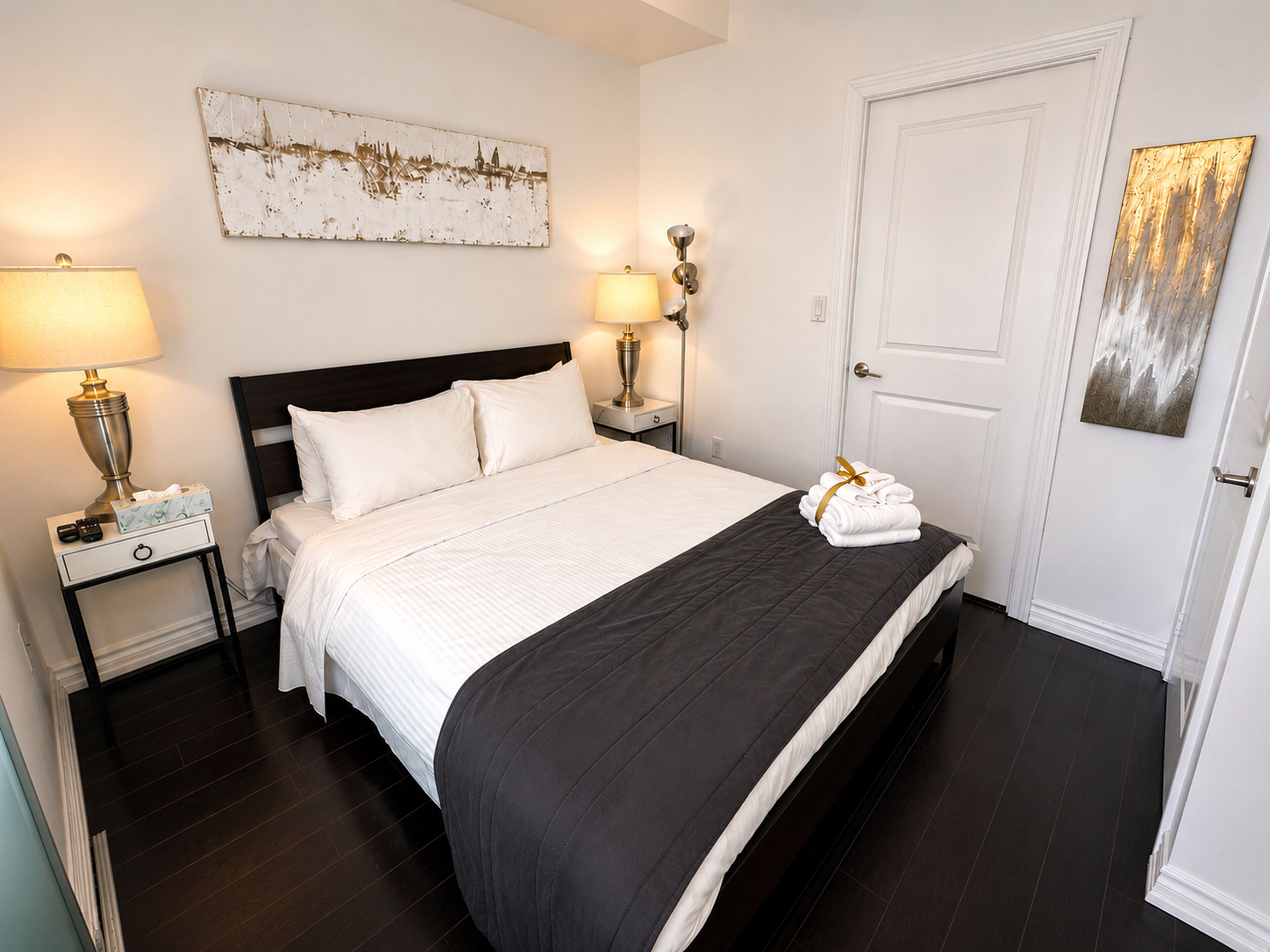 A neatly made bed with white bedding and a black accent, a nightstand with a lamp, a door with a golden handle, and a wall-mounted artwork.