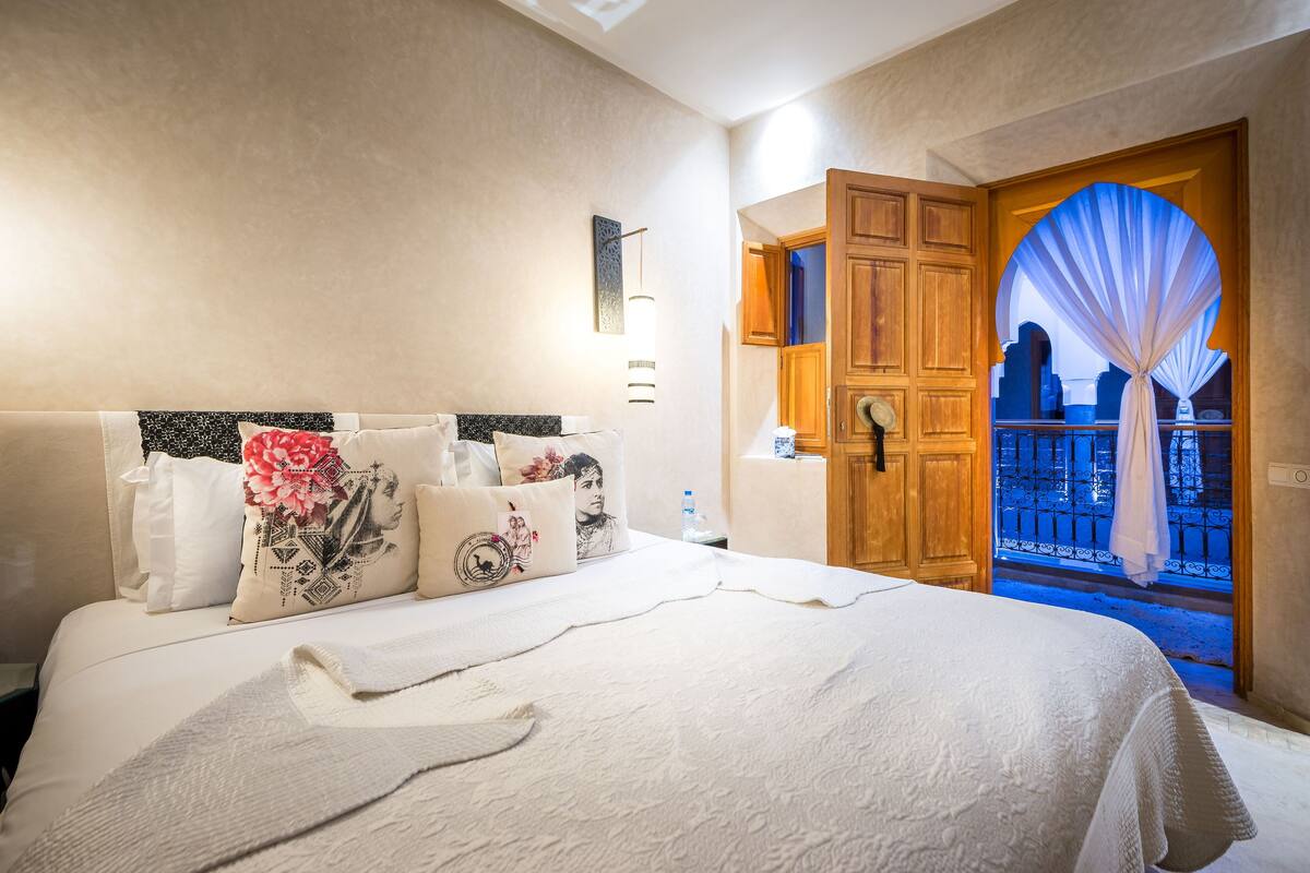 A hotel room with a large bed, a wooden wardrobe, a nightstand, and a window with a view of the outside.