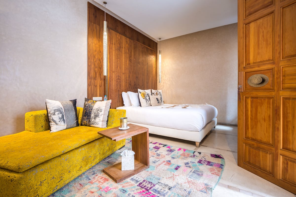 A bedroom with a yellow sofa, a white bed, a wooden wardrobe, and a colorful rug.