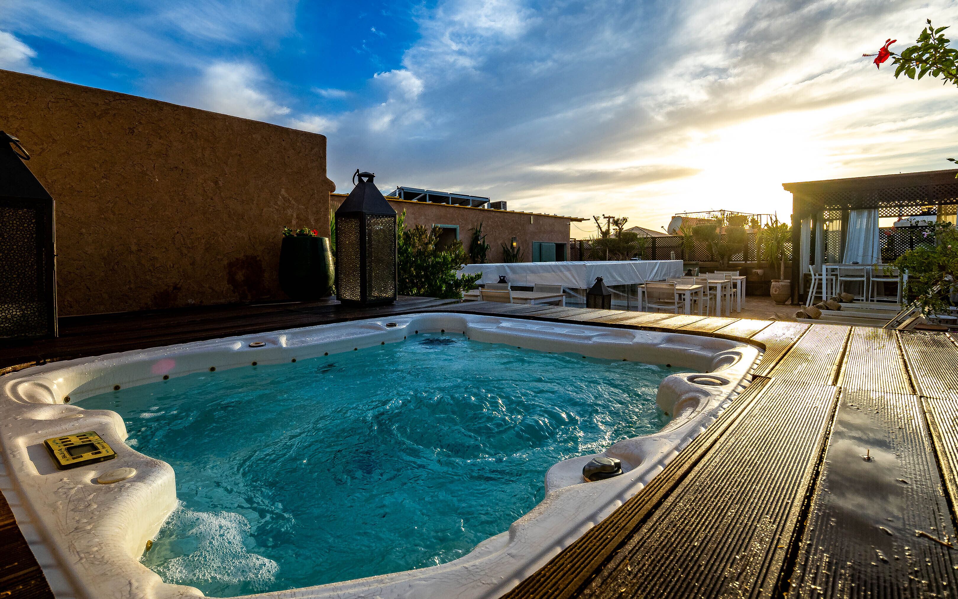 A hot tub with a wooden deck, a table and chairs, and a pergola with a table underneath.